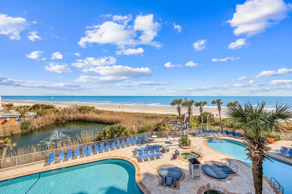 Outdoor pools and blue lounge chairs beside a pond, with the beach and ocean beyond